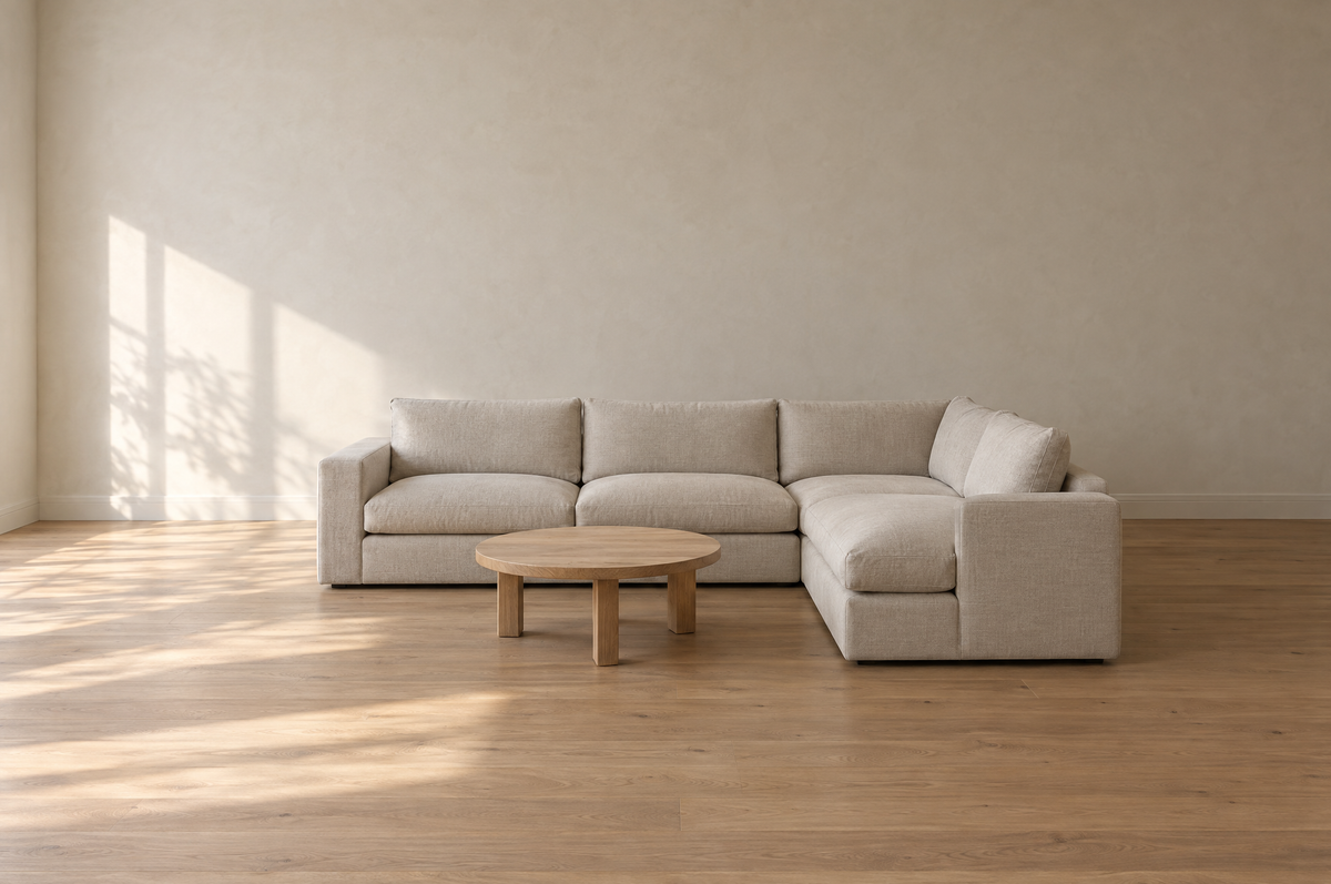 A sunlit living room with a plain oatmeal L-shaped sectional and a round wooden coffee table, surrounded by open floor space.