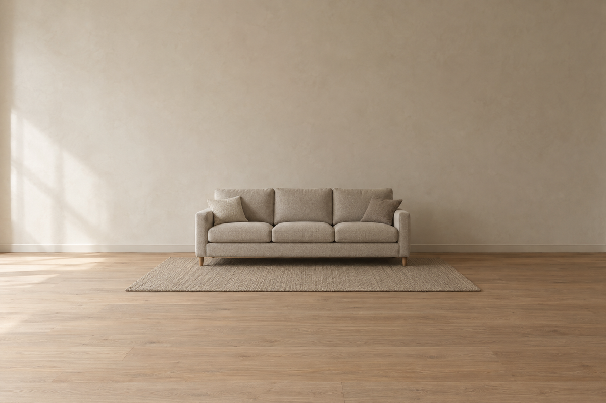 A quiet, empty living room in warm natural light with a neutral oatmeal three-seat fabric sofa centered on a flat-weave rug against a matte plaster wall