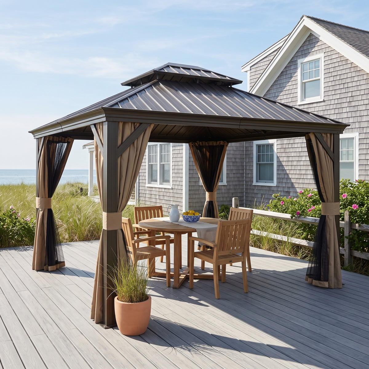 Brown double roof hardtop gazebo with tan curtains over a teak dining set on a gray deck by a shingled beach house