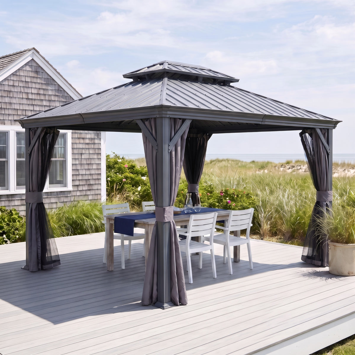 Gray 10x12 hardtop gazebo with tied back curtains over a white dining set on a coastal deck by a shingled house