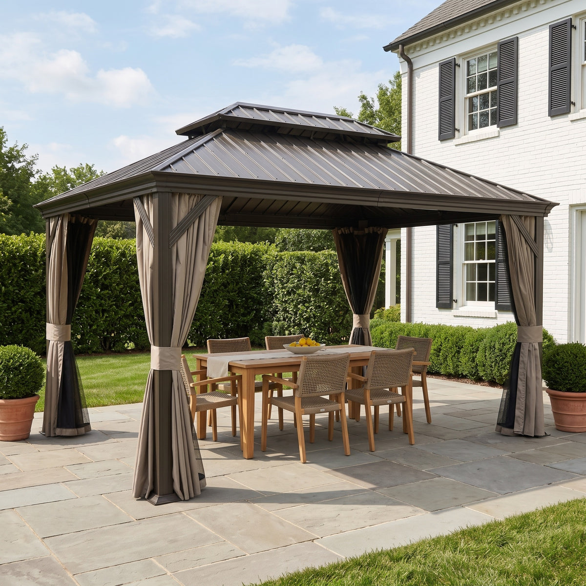 Brown 10x14 hardtop gazebo with tied back curtains over a teak dining table on a stone patio beside a white brick house