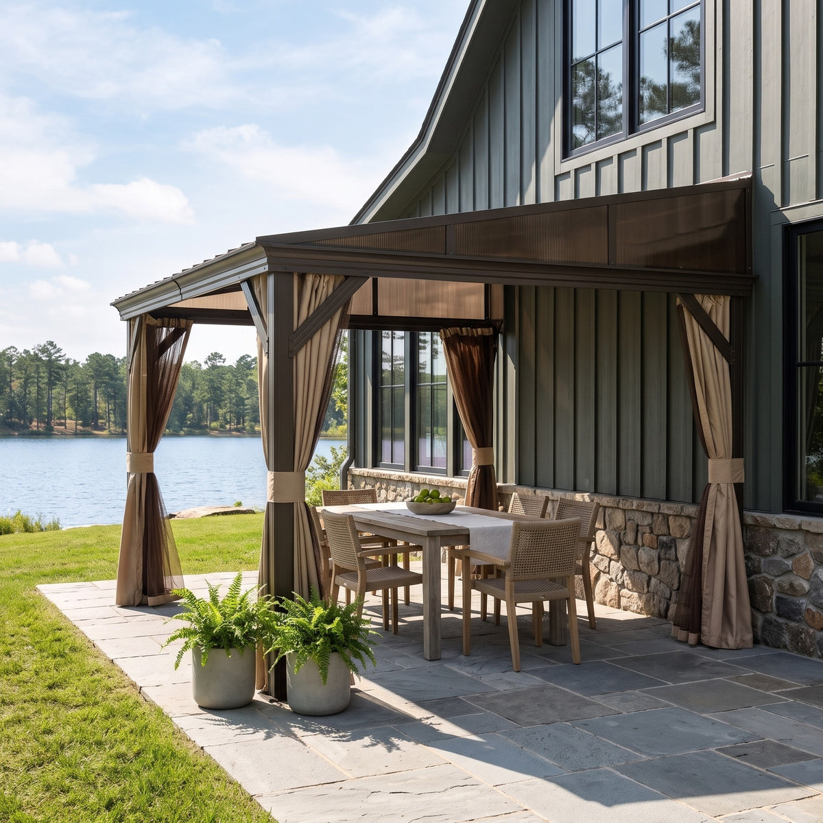 Dark brown steel lean-to hardtop gazebo against a green house wall over a dining table on a stone patio by a lake