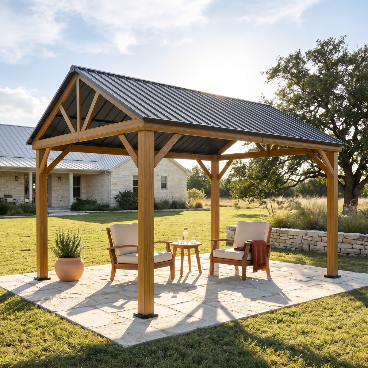 Wood-look aluminum 12x14 hardtop gazebo with gray steel gable roof over two lounge chairs on a stone patio by a lawn