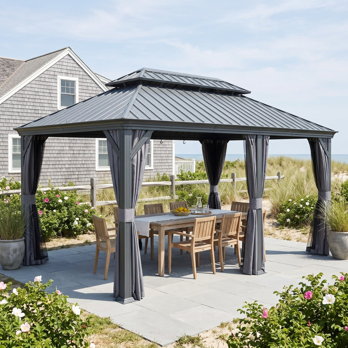 Gray 12x18 hardtop gazebo with tied back curtains over a teak dining table on a stone patio by the beach