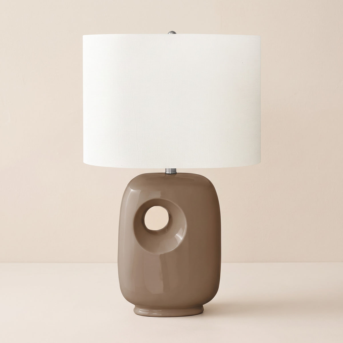 Brown glossy ceramic table lamp with circular cut-out base and ivory linen-look drum shade on an ivory backdrop