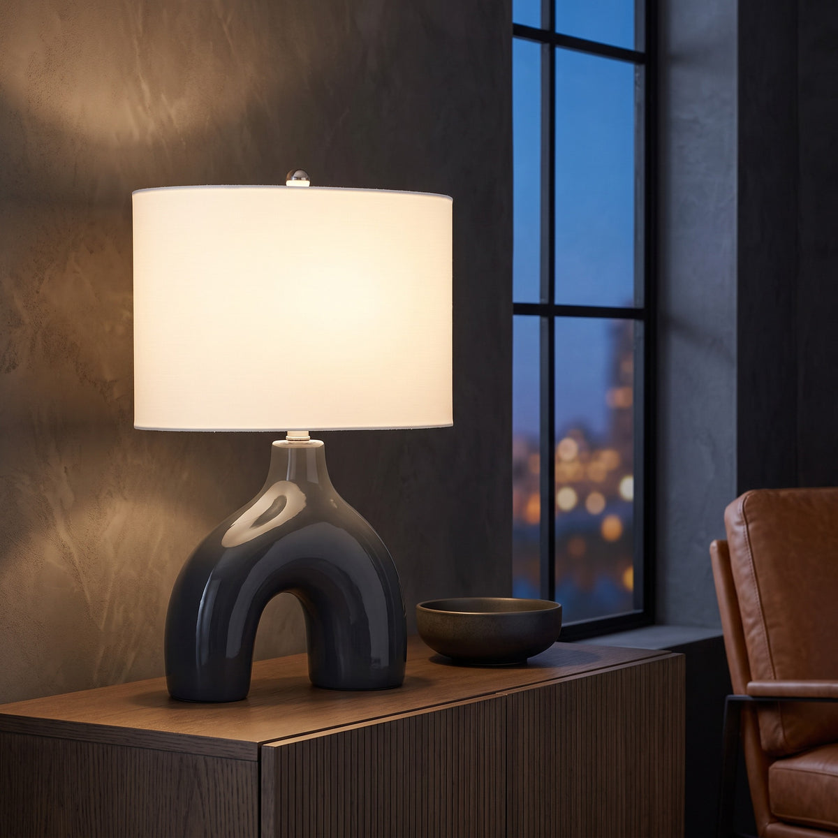 Grey ceramic arch table lamp with ivory drum shade lit on a walnut sideboard by a window at dusk