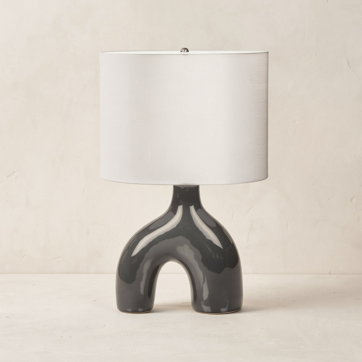 Dark grey glossy ceramic arch table lamp with white linen-look drum shade on an ivory plaster backdrop