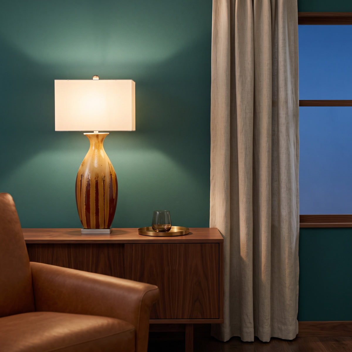 Ceramic table lamp with red and ochre drip glaze and ivory rectangular shade, lit on a walnut credenza by a teal wall