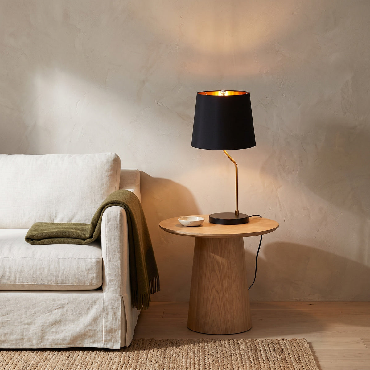 Black table lamp with brass stem and gold-lined shade, lit, on an oak side table beside a white linen sofa