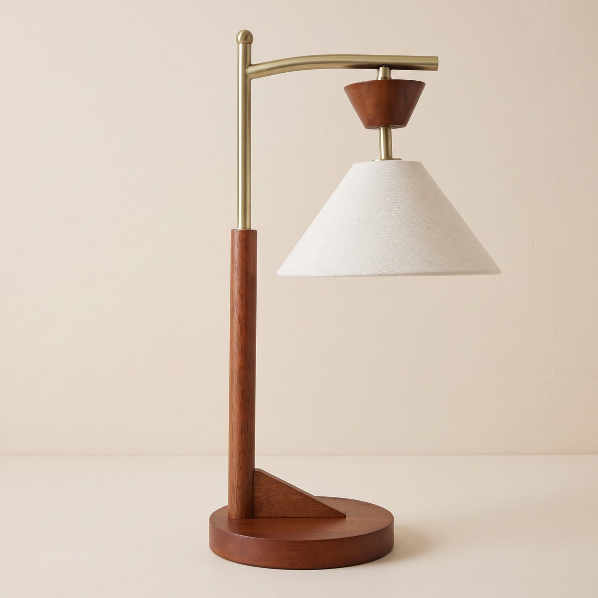 Brown wood table lamp with brass arm and off-white linen-look cone shade on a warm ivory backdrop