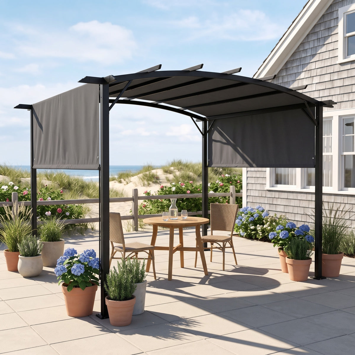 Gray metal pergola with retractable shades over a bistro table on a sunny coastal patio beside a shingled house