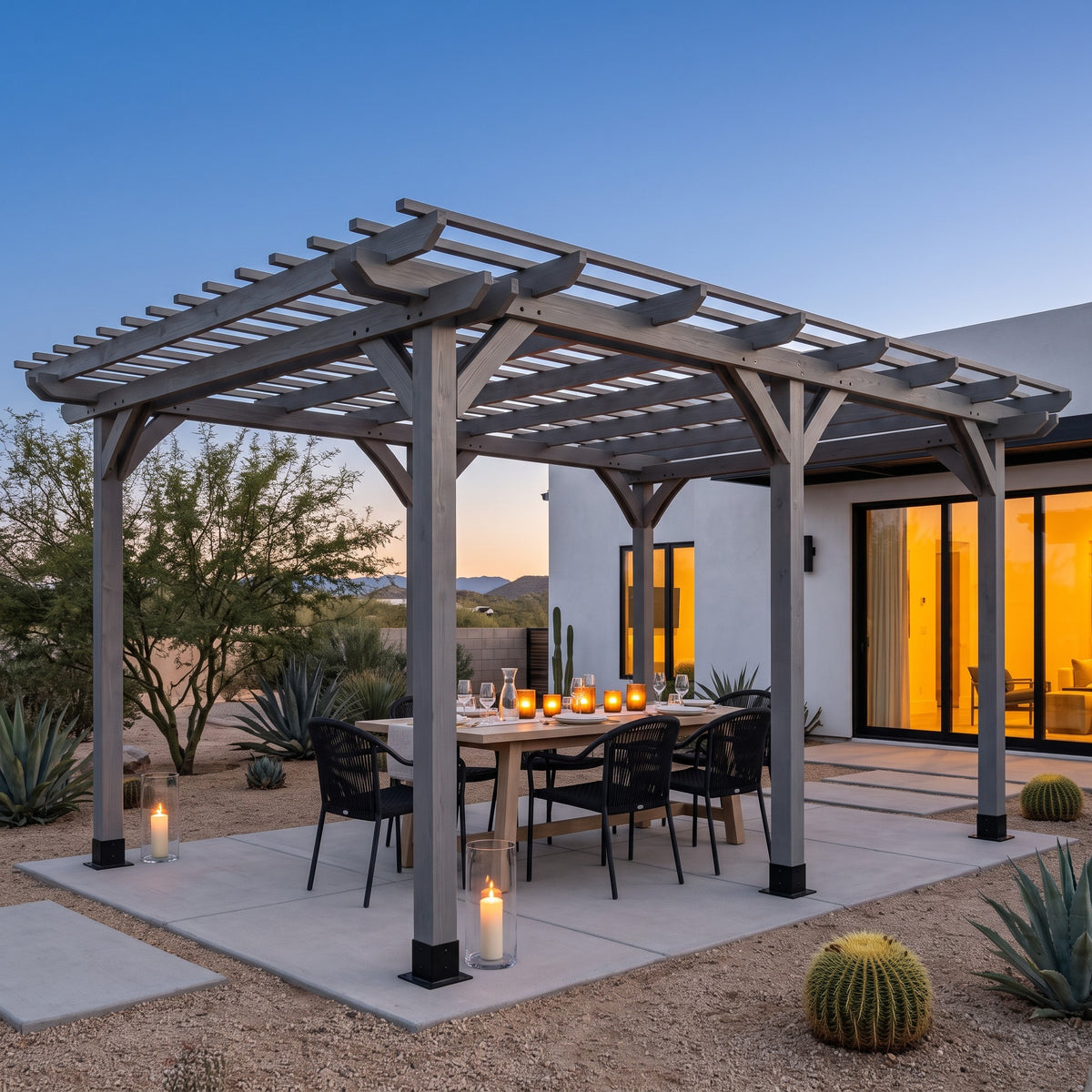 Gray wood pergola over a candlelit dining table on a concrete patio beside a desert home at dusk