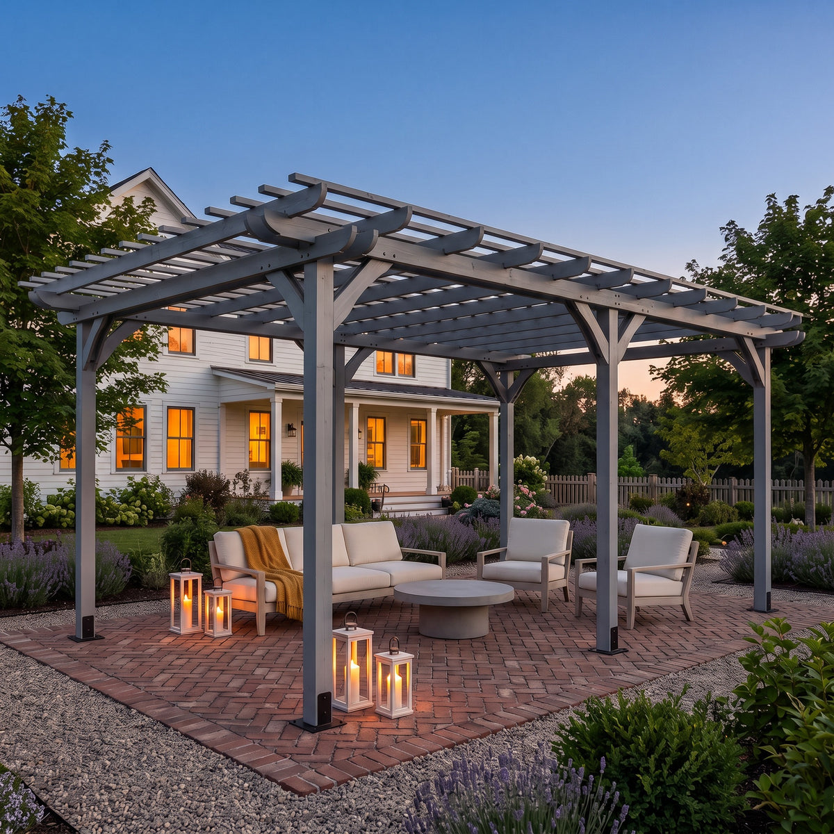 Gray wood pergola over a sofa, armchairs and candle lanterns on a brick patio beside a farmhouse at dusk