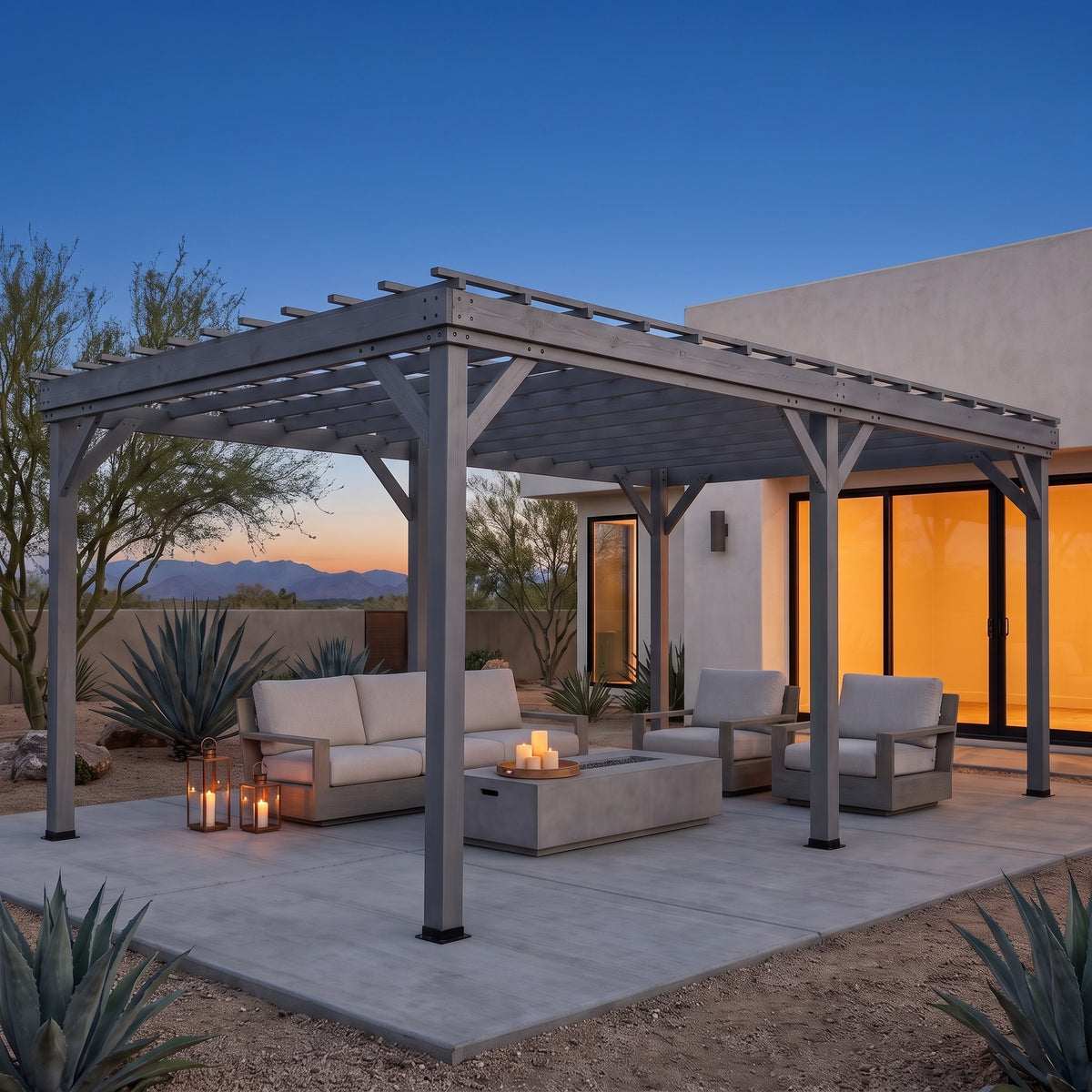 Gray wood pergola over an outdoor sofa and lounge chairs on a concrete patio at dusk with lanterns and candles