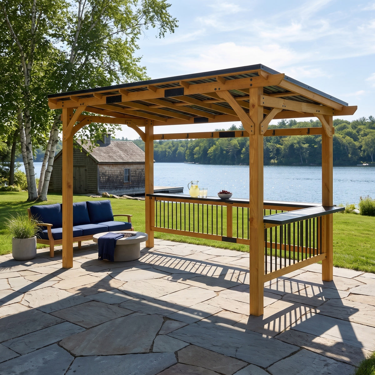 Natural wood gazebo with black metal pent roof and bar shelves on a stone patio beside a lake