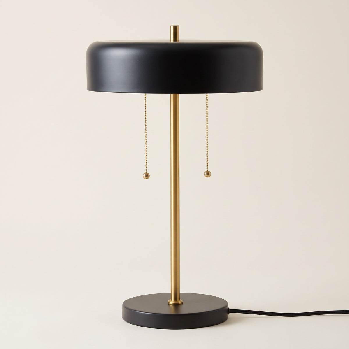 Black metal table lamp with brass stem, drum shade and two pull chains on an ivory backdrop