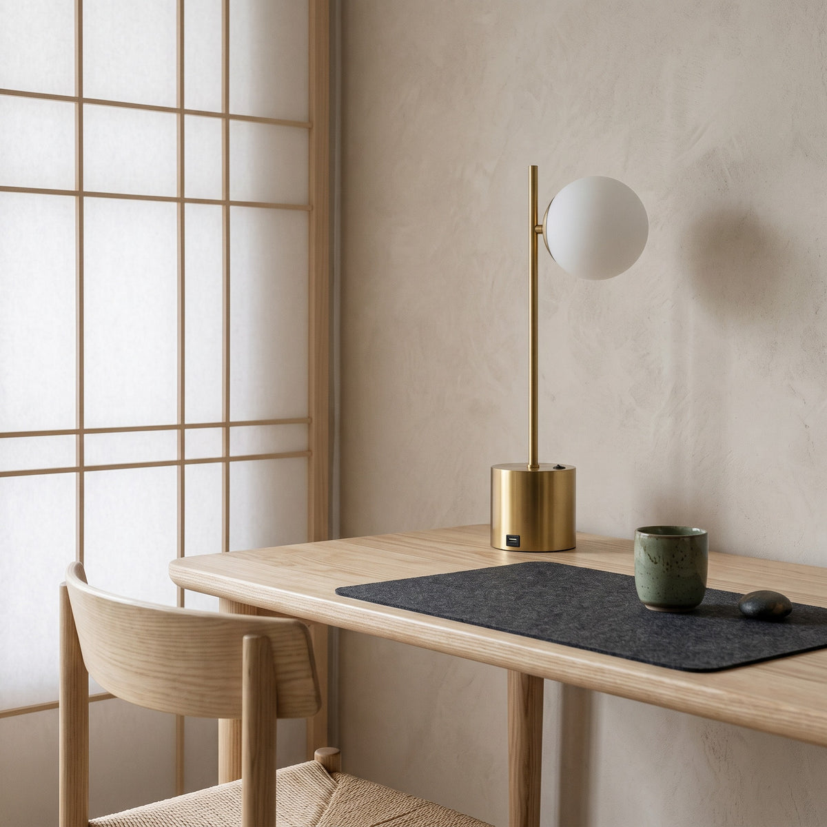 Brass table lamp with white glass globe shade on a light ash desk beside a paper screen window