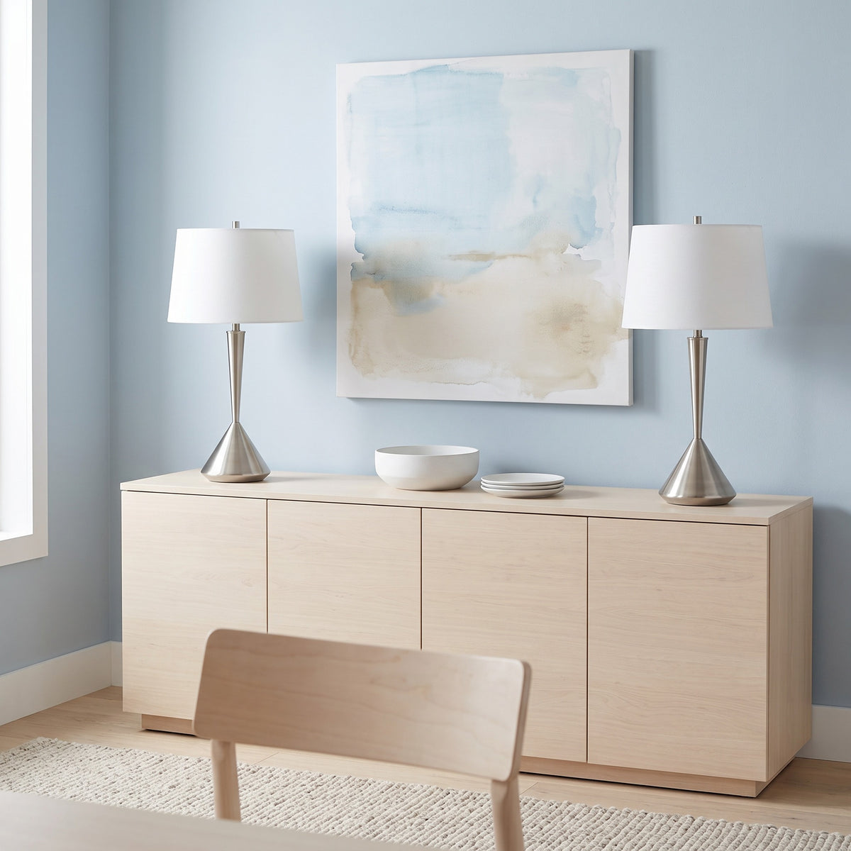 Pair of brushed nickel table lamps with white linen shades on a light maple sideboard against a blue wall