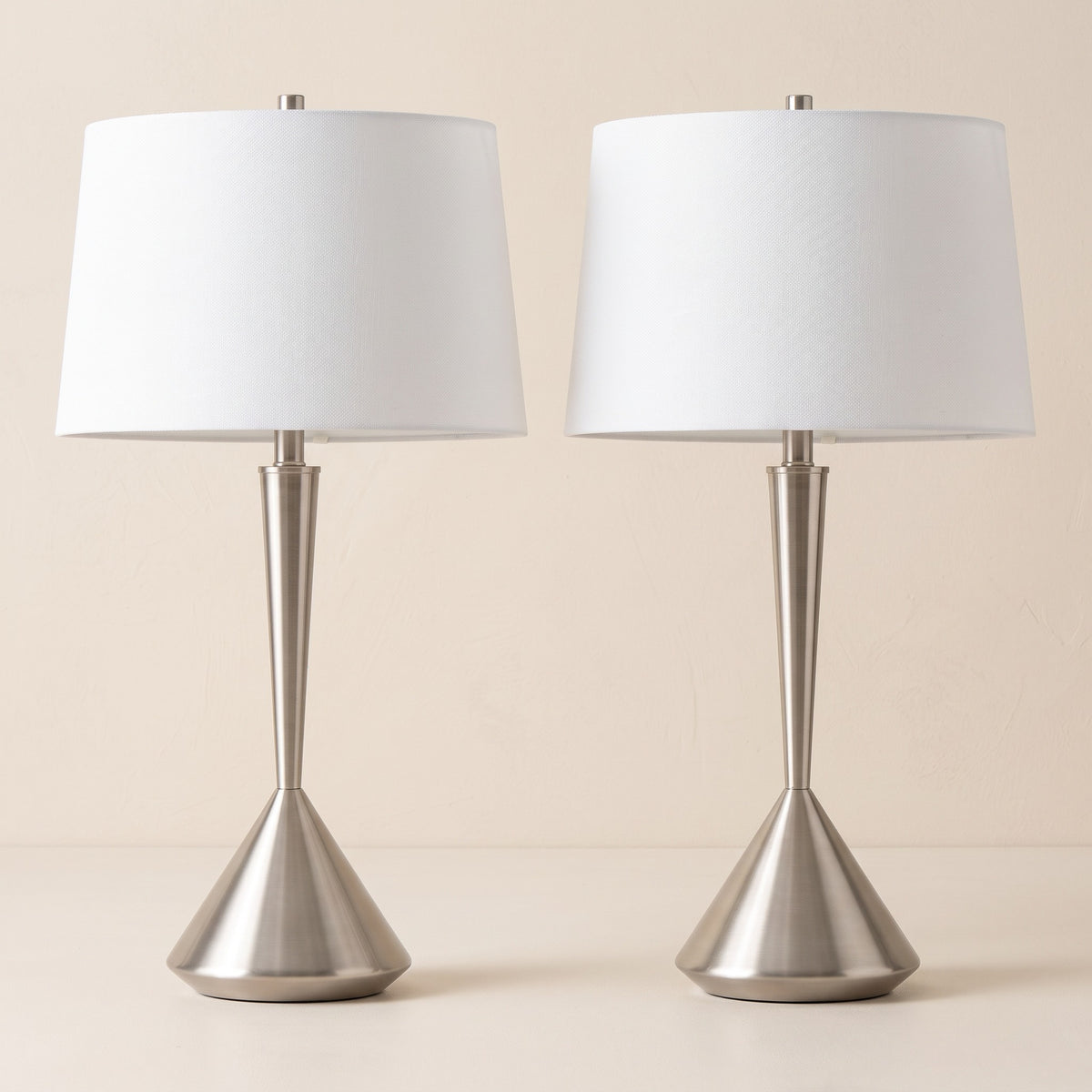 Two brushed nickel table lamps with white tapered linen shades on a warm ivory backdrop