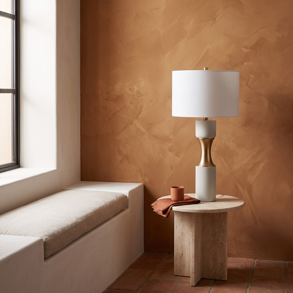 White cement table lamp with brass finish waist and white drum shade on a travertine side table