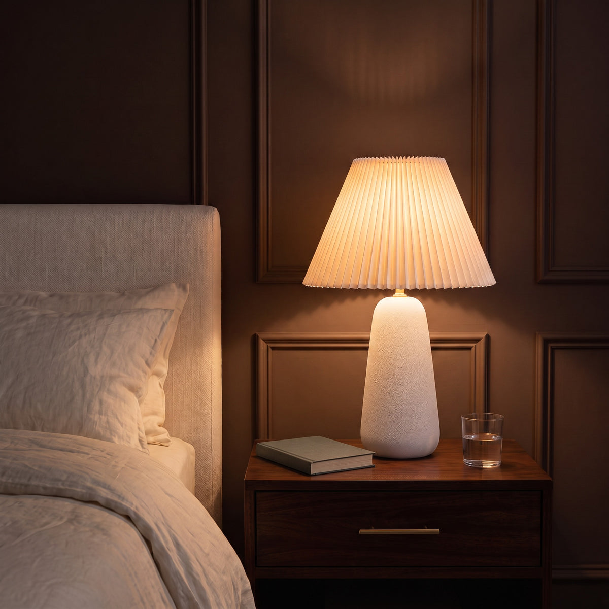 White textured ceramic table lamp with pleated white shade, lit, on a walnut nightstand beside a bed
