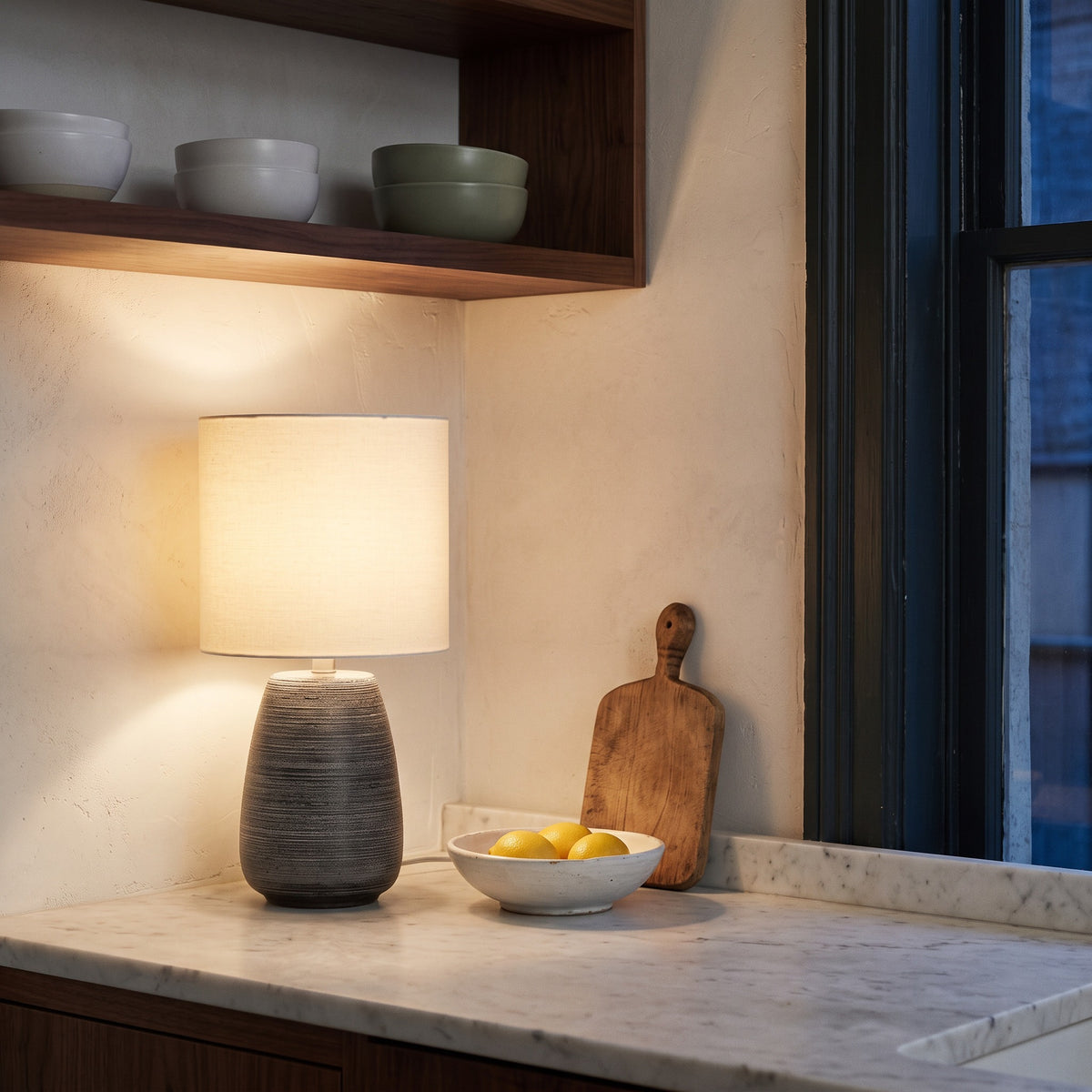 Grey ribbed ceramic table lamp with ivory linen drum shade, lit on a marble kitchen counter beside a bowl of lemons