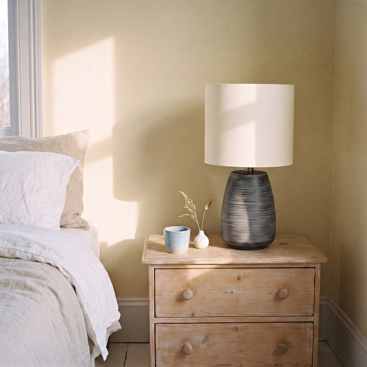 Grey ribbed ceramic table lamp with ivory drum shade on a pine nightstand beside a bed in morning light