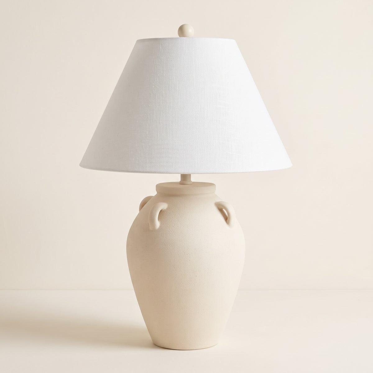 Beige ceramic jug table lamp with handles and white linen-look empire shade on an ivory backdrop