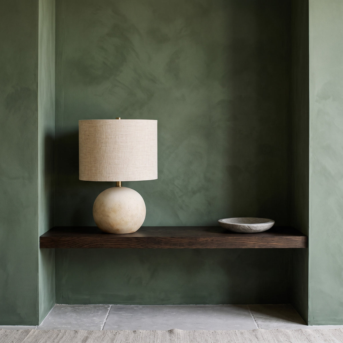 Beige concrete ball table lamp with linen drum shade on a dark oak shelf against a green plaster wall