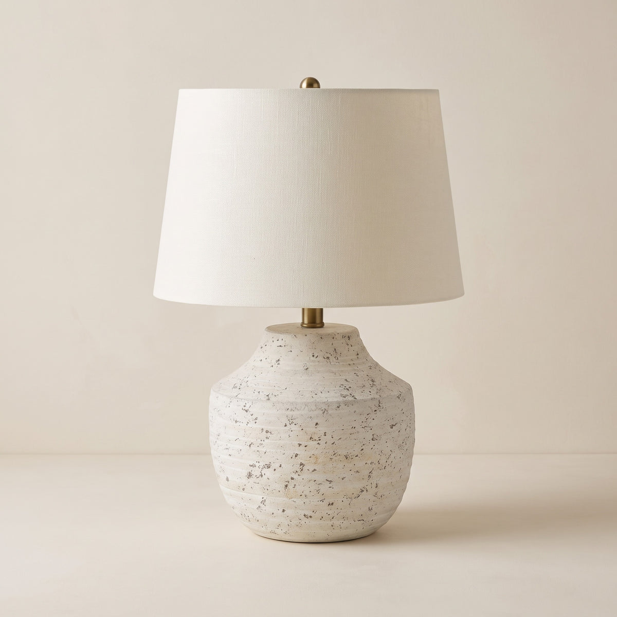 Cream concrete table lamp with ribbed urn base, brass neck and ivory linen drum shade on an off-white backdrop