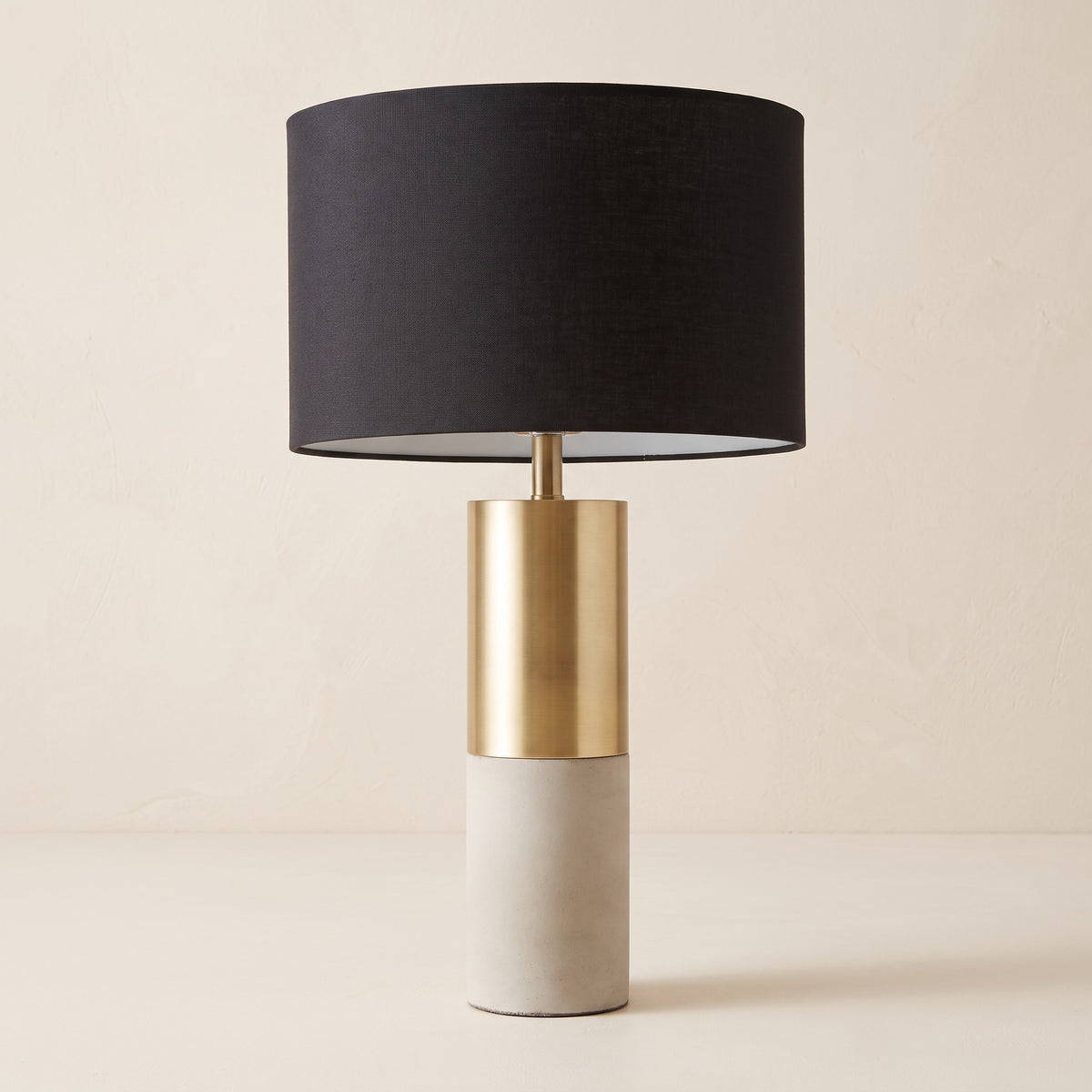 Table lamp with brass and concrete column base and black drum shade on an ivory plaster backdrop