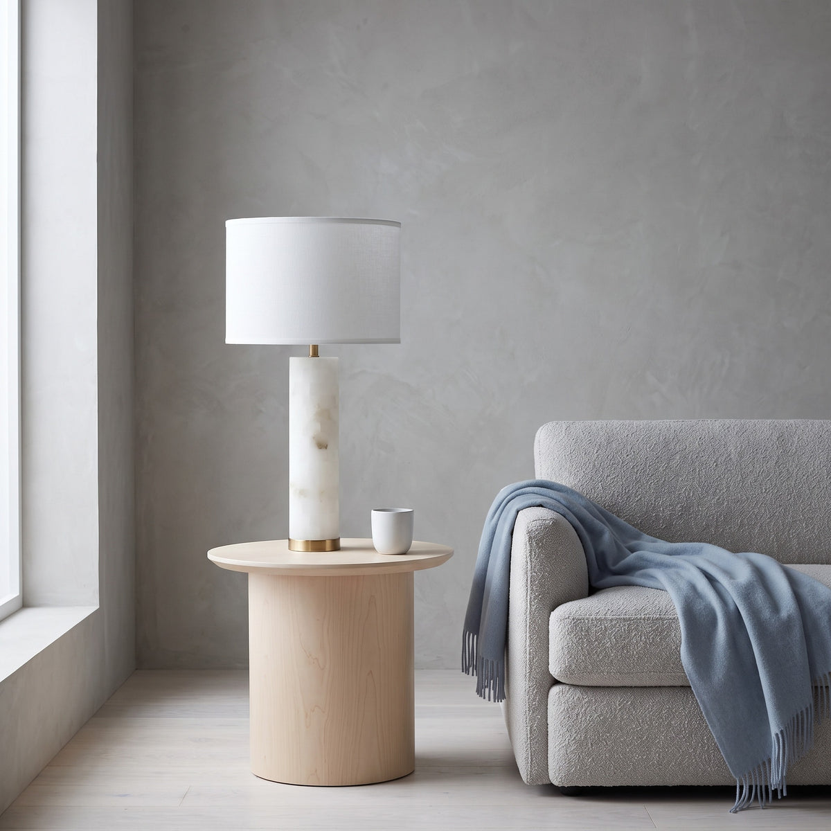 White alabaster table lamp with gold base and white linen drum shade on a maple side table by a gray sofa