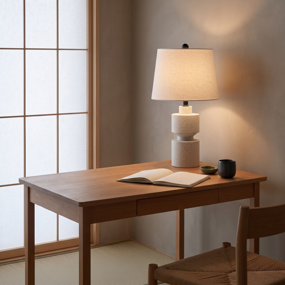 Speckled beige resin table lamp with white linen shade lit on a wood desk beside a paper screen window