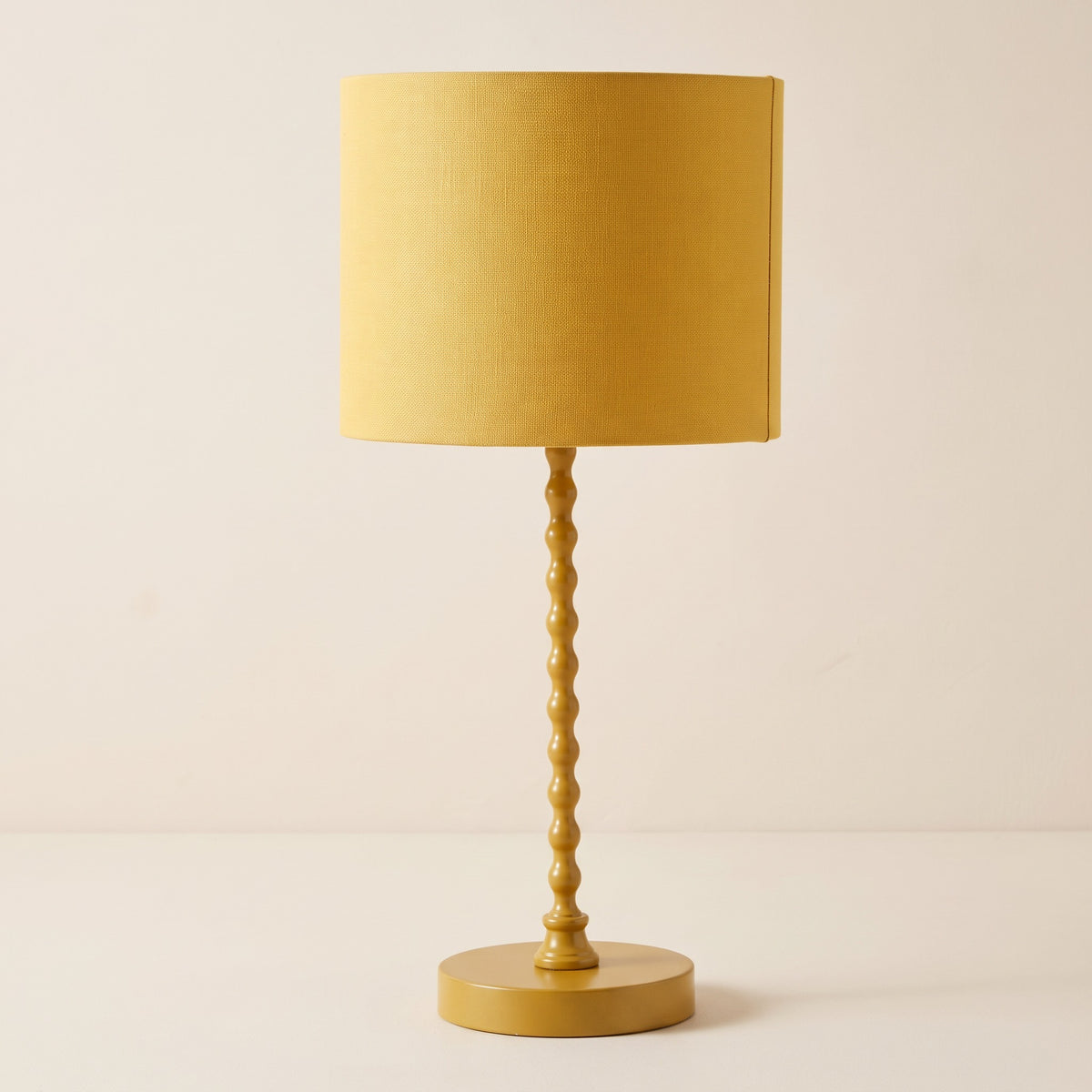 Yellow table lamp with beaded metal stem and linen-look drum shade on a warm ivory backdrop