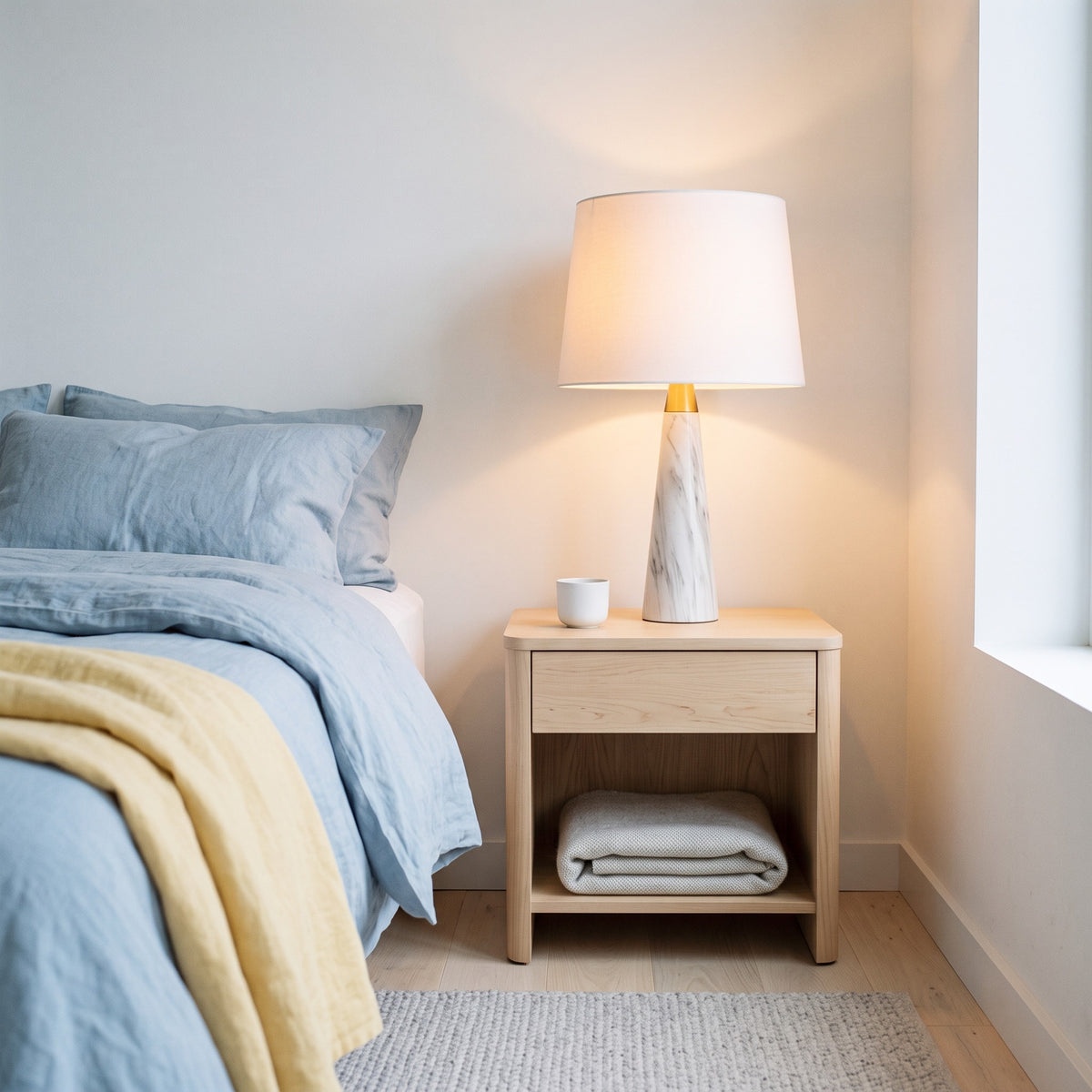 White marble-look table lamp with brass collar and white fabric shade, lit on a light wood nightstand by a bed