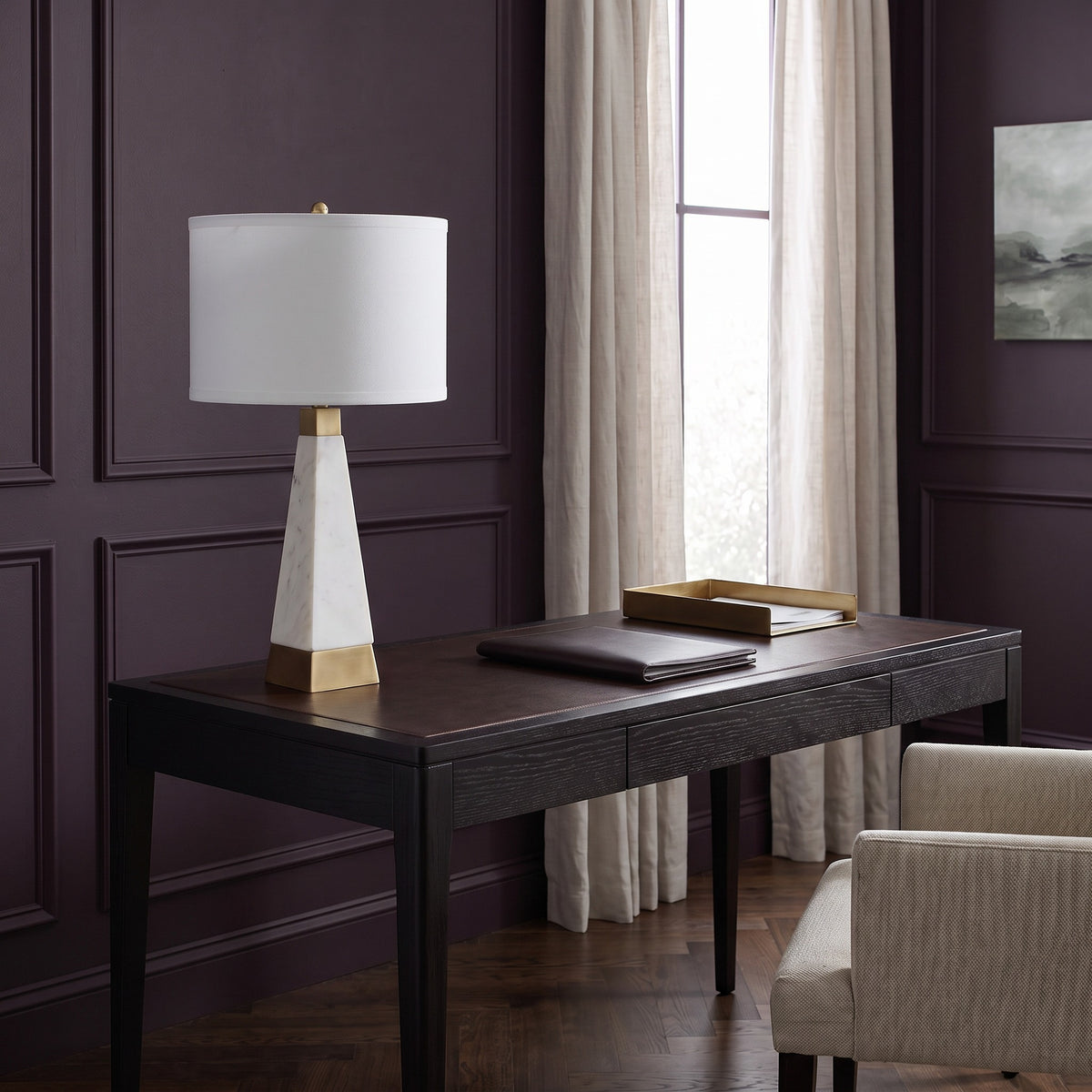 White marble table lamp with brass finish accents and white drum shade on a dark wood desk by a window