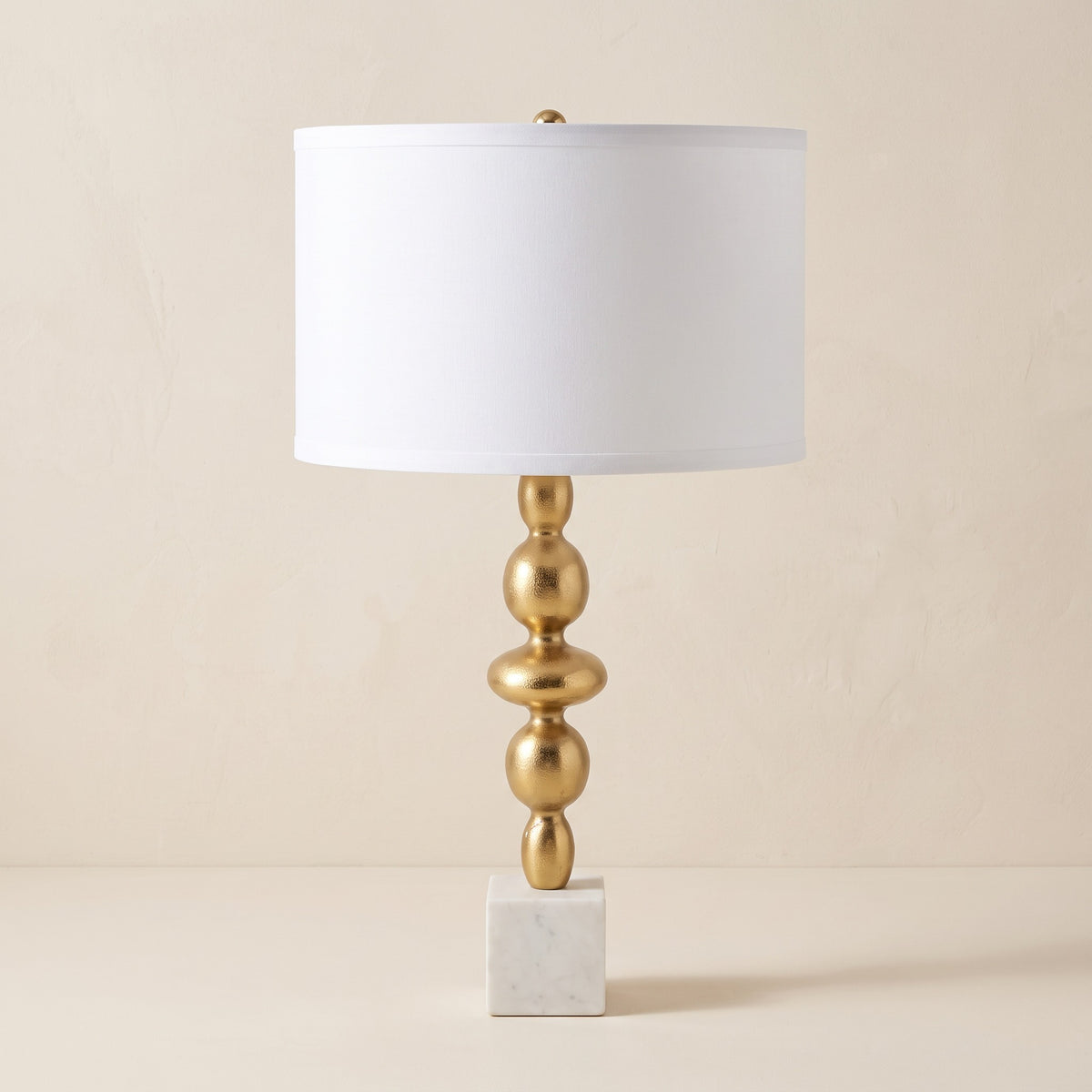 Gold table lamp with stacked orb stem, white marble cube base and white drum shade on an ivory backdrop