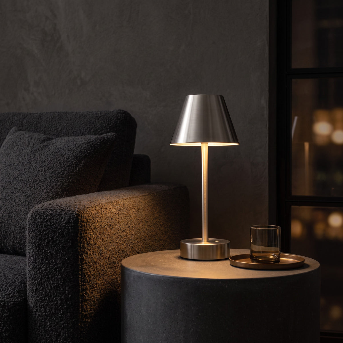 Brushed nickel cordless table lamp glowing on a round black side table beside a charcoal boucle sofa