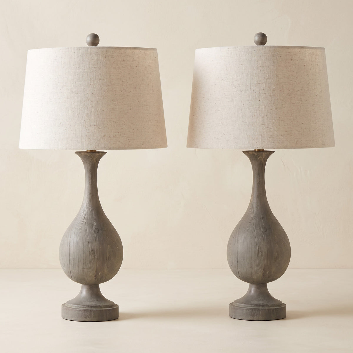 Pair of gray wood-look table lamps with linen drum shades and brass neck accents on an ivory plaster backdrop