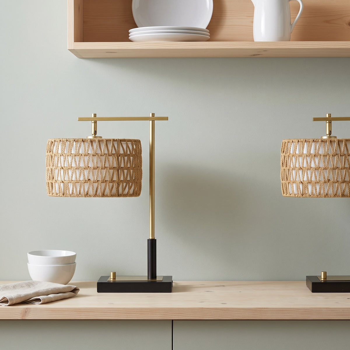 Two woven rope table lamps with gold arms and black bases on a light wood counter below an open shelf