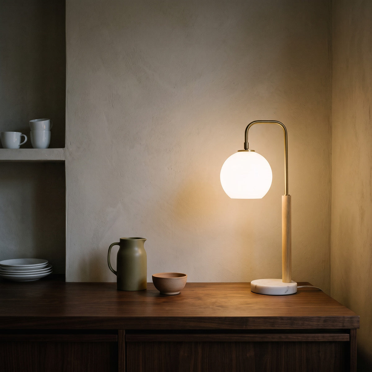 White globe table lamp with brass finish arm, light wood stem and marble base lit on a walnut sideboard