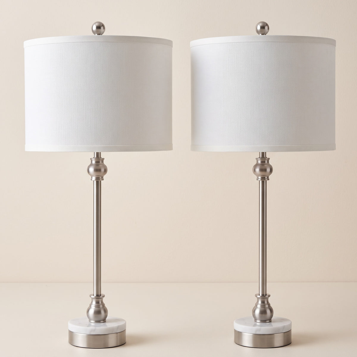 Pair of nickel table lamps with white marble bases and white linen drum shades on an ivory backdrop
