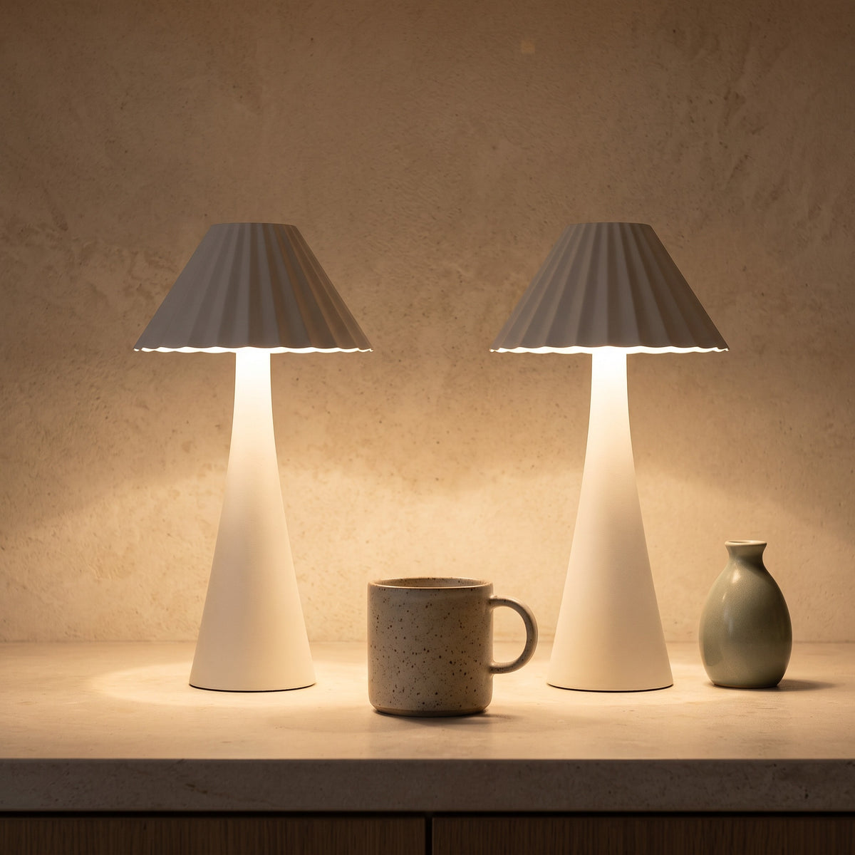 Two white cordless table lamps with pleated shades glowing on a stone counter beside a speckled mug and a sage vase