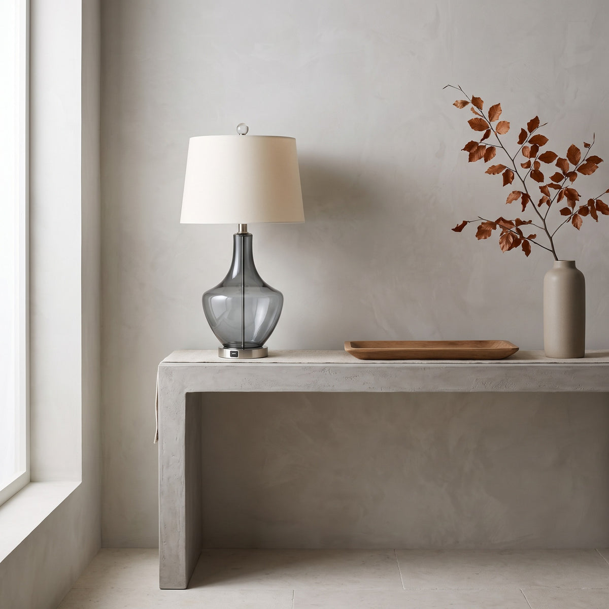 Grey glass table lamp with white linen-look shade and USB port base on a plaster console by a window