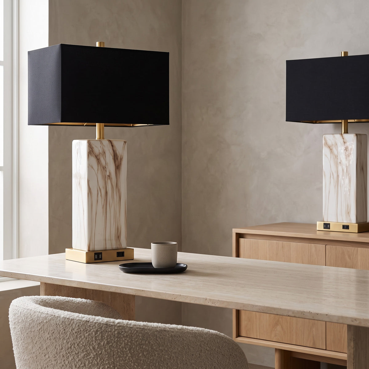Two white marble-look table lamps with black rectangular shades and gold bases on a travertine desk and oak sideboard