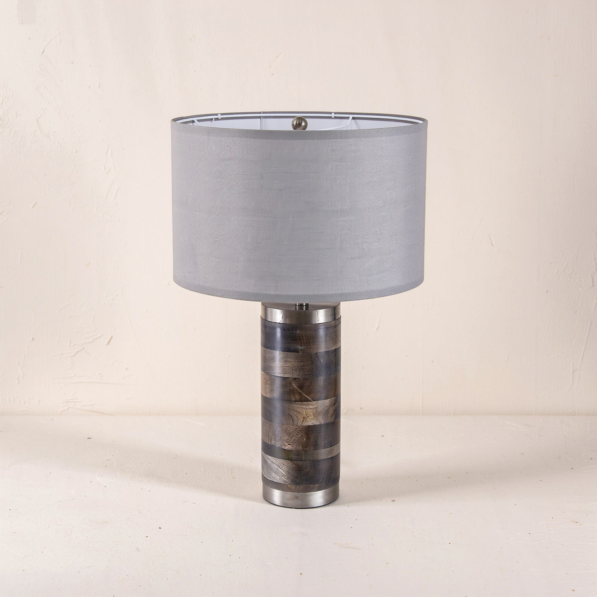 Table lamp with banded gray mango wood base, brushed metal trim and gray drum shade on an ivory plaster backdrop