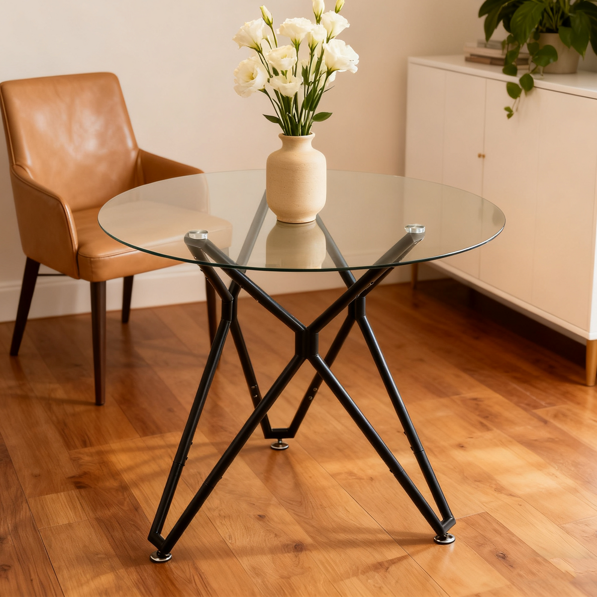 Round glass dining table with a black base and a tan leather chair, ceramic vase of white flowers on top