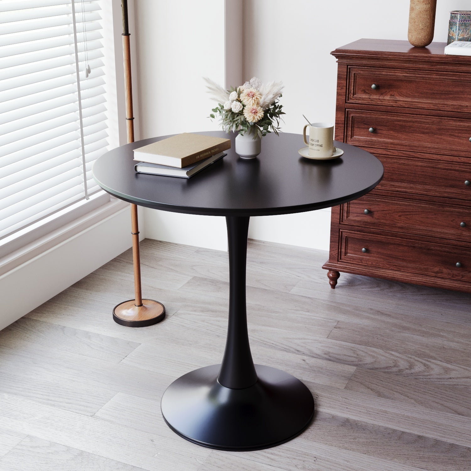 Black round tulip dining table styled with books, mug, and flowers by a window beside a wood dresser