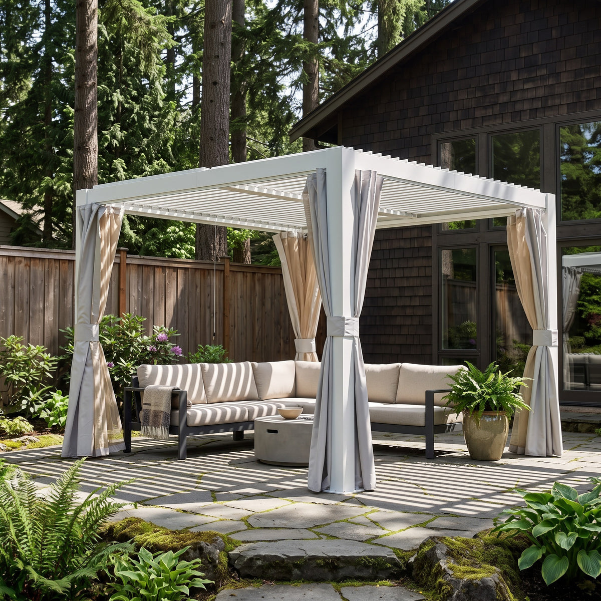 White aluminum louvered pergola with tied back curtains over a beige sectional on a stone patio in a wooded backyard