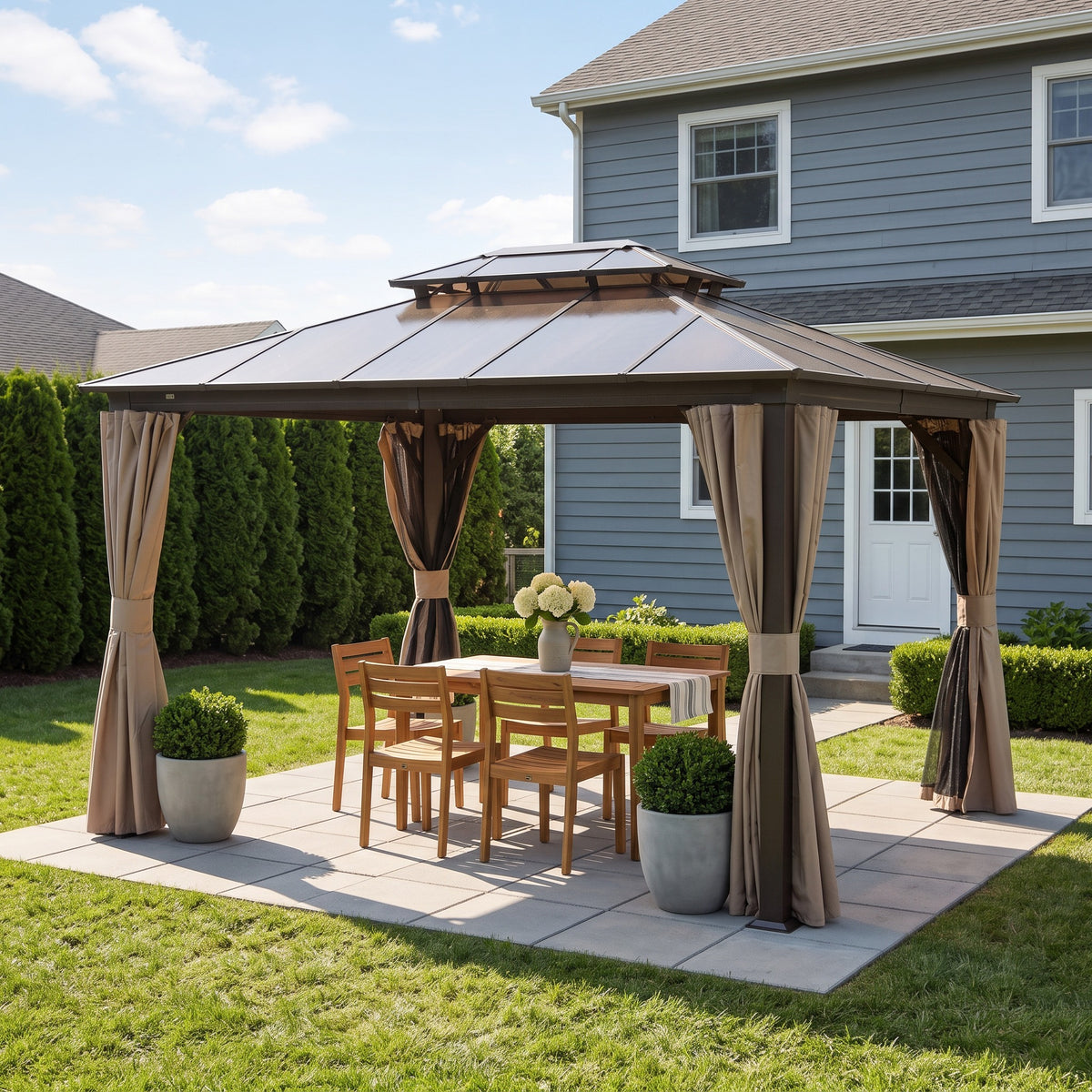 Brown aluminum double roof gazebo with tied back beige curtains over a wood dining set on a paver patio by a gray house